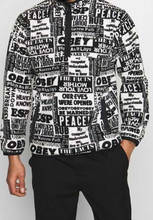 Pull polaire noir et blanc avec des blocs de texte audacieux comprenant "OBEY", "PEACE" et des phrases, porté avec un pantalon noir.