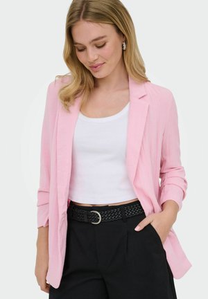 Kobieta ubrana w jasnoróżową marynarkę, biały krótki top, czarne spodnie z plecionym paskiem i perłowe kolczyki, pozująca z jedną ręką w kieszeni.