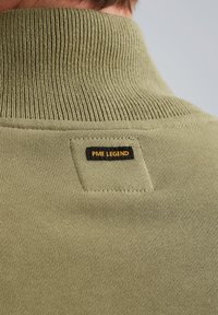 Olijfgroene trui met hoge kraag en ribbelstructuur. Heeft een zwart label met "PME LEGEND" in gele stiksels op de rug.