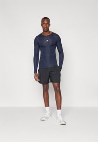 Jūrinės sportinės ilgomis rankovėmis viršus su apvaliu iškirpimu, derinamas su juodais šortais. Baltas logotipas ant krūtinės. Modelis vilki baltus sportbačius ir kojines.