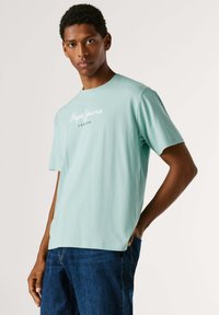 Μπλουζάκι κοντομάνικο σε μέντα πράσινο χρώμα, κατασκευασμένο από βαμβάκι, με στρογγυλή λαιμόκοψη. Διαθέτει λευκό λογότυπο «Pepe Jeans» τυπωμένο στο στήθος. Φορεμένο με μπλε τζιν παντελόνι.