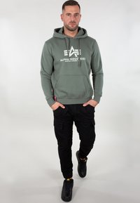 Felpa verde con logo bianco, tasca a marsupio e cappuccio con stringhe. Abbinata a pantaloni cargo neri e sneakers nere con dettagli gialli.