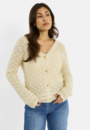 Femme portant un cardigan en tricot crochet beige à boutons sur un haut blanc et un jean bleu, debout les mains près des poches, regardant ailleurs.
