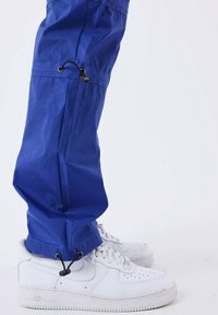 Blauwe nylon broek met verstelbare touwen aan de manchetten. Draagt met witte sneakers die een gestructureerd, sportief ontwerp tonen.