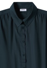 Chemise boutonnée à col vert foncé avec une étiquette de marque « Cecil », tissu lisse et légers fronces aux coutures des épaules.