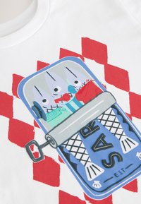 Fehér pamutpóló, mely színes grafikai mintával díszített, konzervezett halakat ábrázol különböző színekben, piros kockás háttérrel. Előnyös szabású és kerek nyakú.