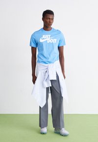 T-shirt de algodão azul com o texto branco "FAÇA ACONTECER", calças plissadas cinzentas e uma camisa de botão azul claro amarrada à cintura. Ténis cinzentos.