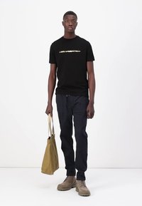 T-shirt en coton noir avec logo doré, pantalon bleu marine, bottes beiges et sac fourre-tout beige. Design simple et décontracté. Fond uni.