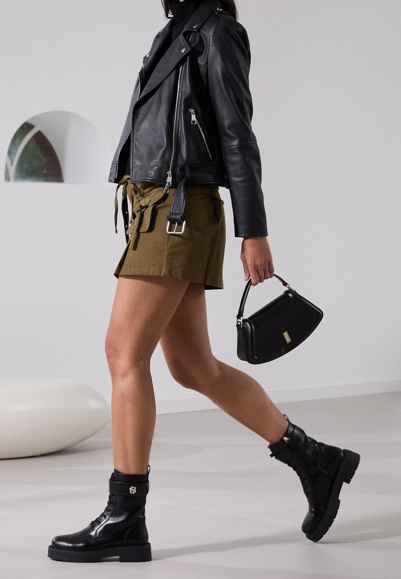 Veste de moto en cuir noir, shorts vert olive avec ceinture, bottines noires, petit sac à main noir ; design moderne, textures élégantes.
