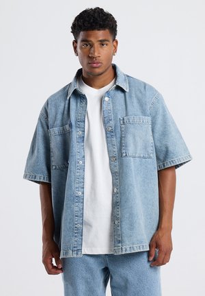 Lichtblauw denim overhemd met korte mouwen, twee voorzakken, knoopsluiting en een ontspannen pasvorm, gedragen over een wit T-shirt.
