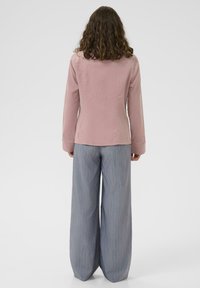 Žena s kosom do ramena koja nosi ružičasti džemper s dugim rukavima i široke plave pantalone s prugicama, stoji okrenuta leđima.