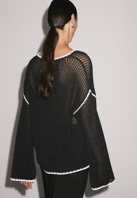 Femme aux longs cheveux noirs en queue de cheval basse, portant un pull en mesh noir large avec des bordures blanches, vue de dos sur un fond uni.