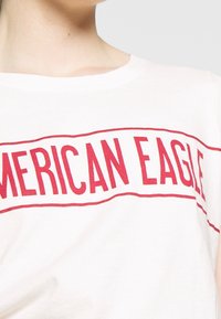 Weißes Baumwoll-T-Shirt mit rotem "AMERICAN EAGLE"-Text, weitem Ausschnitt und lockerem Schnitt. Minimalistisches Design ohne zusätzliche Muster oder Akzente.