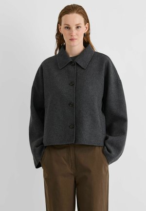 Femme aux cheveux mouillés portant une veste gris foncé boutonnée avec un large col et un pantalon marron taille haute, les mains dans les poches, debout devant un fond uni.