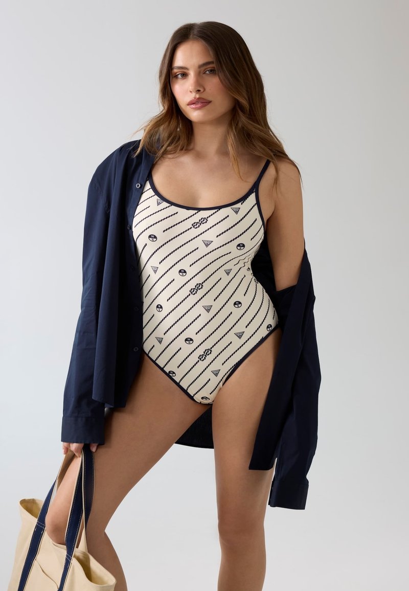 Jeune femme portant un maillot de bain blanc à motifs et une chemise bleu marine ouverte, tenant un sac fourre-tout beige sur un fond clair uni.