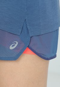 Shorts de course en filet bleu avec une fente latérale superposés à des shorts de compression corail vif sur la cuisse d'une personne.