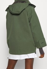 Veste à capuche verte avec une coupe décontractée, dotée d'un tissu texturé et d'accents de poignets en velcro. Présente un design minimaliste avec une grande poche arrière.