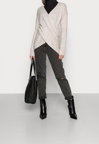 Femme portant un pull enveloppant beige clair sur un turtleneck noir, un jean gris foncé déchiré, des bottes noires à bout pointu, tenant un sac à main en cuir noir.