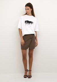 Tricou alb supradimensionat cu un imprimeu abstract negru, asociat cu o fustă înfășurată maro cu detalii cu buton și sandale cu toc negre.
