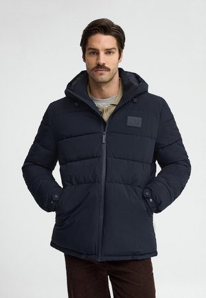 Uomo con baffi che indossa una giacca imbottita nera con cappuccio e le mani nelle tasche, in piedi davanti a uno sfondo chiaro e uniforme.