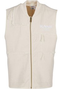 Beige ermeløs vest med glidelås, med frynset detaljer, to frontlommer og broderte tekster på brystet.