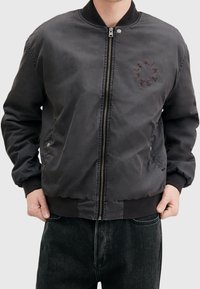 Hombre vistiendo una chaqueta bomber negra con cremallera frontal, puños y.do con elástico, y un logotipo circular en el lado izquierdo del pecho. Manos parcialmente en los bolsillos.