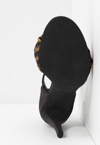 Chaud devant à talons noirs avec une semelle à motif de logo et un dessus en tissu rehaussé d'or, reposant contre un mur blanc.