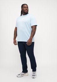 Light blue short-sleeve t-shirt with a white "Belstaff" logo, paired with dark blue jeans and gray and white sneakers on a neutral background.