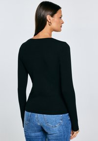 Naine, kellel on pikad tumedad juuksed, kuldsed rõngakujulised kõrvarõngad, must pika varrukaga särk ja sinised teksapüksid, seisab külje pealt palja tausta vastu.