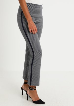 Frau trägt schwarz-weiß gemusterte, gekürzte Hosen mit schwarzem Seitenstreifen und schwarze spitze High Heels mit Knöchelriemen.