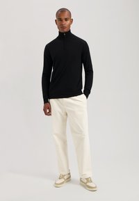 Maglione nero a maniche lunghe con collo a zip abbinato a pantaloni crema chiara e sneakers beige. Design semplice e texture liscia.
