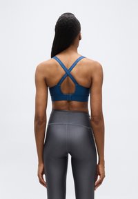 Γυναίκα που φοράει σπορ σουτιέν σκούρου μπλε χρώματος με σταυρωτούς ιμάντες και γκρι κολάν με ψηλή μέση, στέκεται με την πλάτη στραμμένη προς την κάμερα μπροστά σε λευκό φόντο.