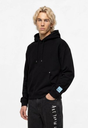 Jeune homme aux cheveux blonds portant un sweat à capuche noir et un pantalon noir avec un texte blanc ""ARE YOU IN"" sur la jambe, debout avec une main dans la poche.