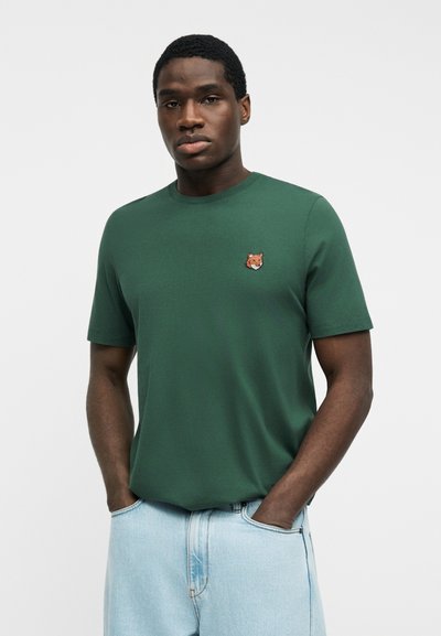 Άνδρας που φοράει σκούρο πράσινο κοντομάνικο πουκάμισο με ένα μικρό έμβλημα αλεπούς στο στήθος και ανοιχτό μπλε τζιν παντελόνι, με τα χέρια στις τσέπες.