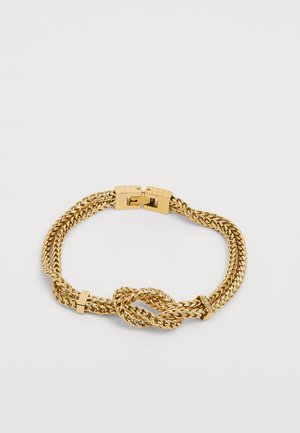 Auksinės spalvos apyrankė, pagaminta iš pintų grandinėlių, su mazgo detale ir užsegimu. Lygus paviršius su blizgiu finišu.