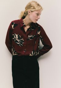 Cămașă călcată bordo cu un model floral negru și crem, cu mâneci lungi și buzunar pe piept. Asortată cu pantaloni negri.