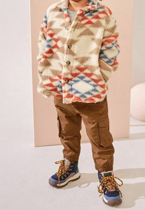 Dziecko ubrane w wzorzysty polarowy kurtkę, brązowe spodnie typu cargo oraz niebieskie buty do trekkingu z żółtymi sznurowadłami, stojące z rękami w kieszeniach.