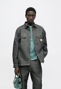 Ciemna jeansowa koszula zapinana na guziki z kontrastowymi białymi szwami, zestawiona z turkusowym t-shirtem i ciemnymi jeansowymi spodniami, trzyma zieloną torbę na ramię.