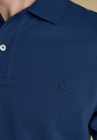 Gros plan d'une personne portant un polo bleu marine avec un petit logo animal brodé près de la poitrine et un bouton blanc près du col.