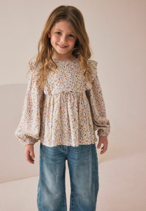 Blumenbluse mit Rüschenärmeln aus hellrosa Stoff, mit einem mehrfarbigen Blumenmuster, kombiniert mit locker sitzenden blauen Jeanshosen.