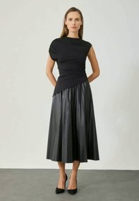 Schwarzes Kleid mit enganliegendem Oberteil und plissierten Lederrock. Glatter, dehnbarer Stoff oben; glänzende, strukturierte Textur unten. Schwarze High Heels.