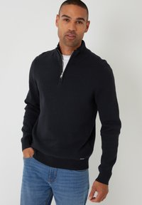 Czarny sweter na zamek wykonany z gładkiej, dzianinowej tkaniny, z wysokim kołnierzem i prążkowanymi mankietami, zestawiony z niebieskimi dżinsami.