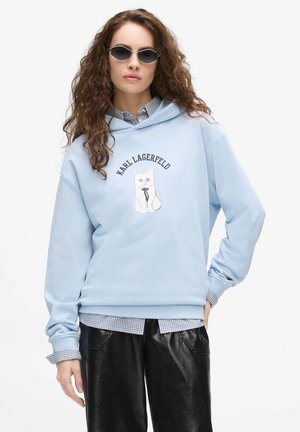 Femme aux longs cheveux bouclés portant des lunettes de soleil, un sweat à capuche bleu clair avec un chat et le texte "Karl Lagerfeld", une chemise à carreaux et un pantalon noir.