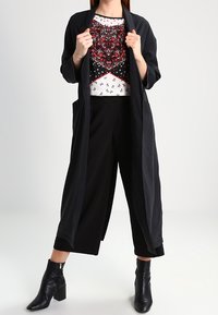 Un lungo cardigan nero sopra una blusa a fantasia, con motivi floreali in rosso e bianco. Abbinato a pantaloni culotteneri larghi e stivali alla caviglia.