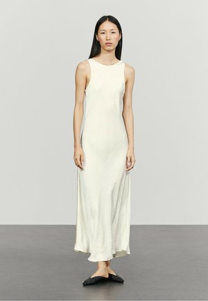 Donna con lunghi capelli scuri che indossa un abito lungo bianco strutturato senza maniche e scarpe nere piatte e appuntite, in piedi davanti a uno sfondo grigio uniforme.