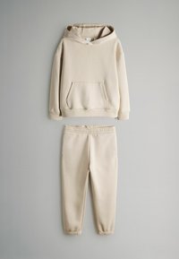 Beige Kapuzenpullover mit Kängurutasche und passender Jogginghose mit elastischem Bund flach auf grauem Hintergrund ausgelegt.