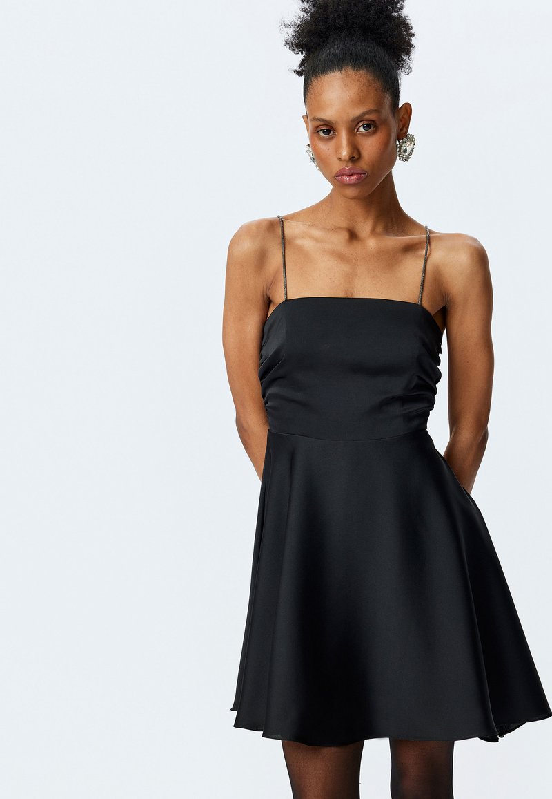 Abito in satin nero con corsetto aderente e gonna a ruota. Presenta sottili spalline regolabili e una leggera arricciatura sul davanti.