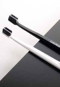 Två CURAPROX-tandborstar, en svart och en vit, har mjuka borst och en stilren ergonomisk design, placerade på en texturerad yta.