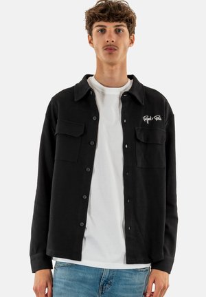 Zwarte button-up shirt van textiel met structuur, voorzien van twee borstzakken en een geborduurd logo. Draagt over een wit t-shirt, gecombineerd met lichte jeans.