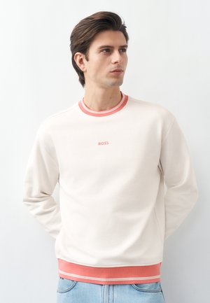 Jaunas vyras su tamsiais plaukais, dėvintis kreminio atspalvio džemperį su koralų spalvos apvadais ir užrašu „BOSS“, derinantis su šviesiai mėlynais džinsais, baltame fone.